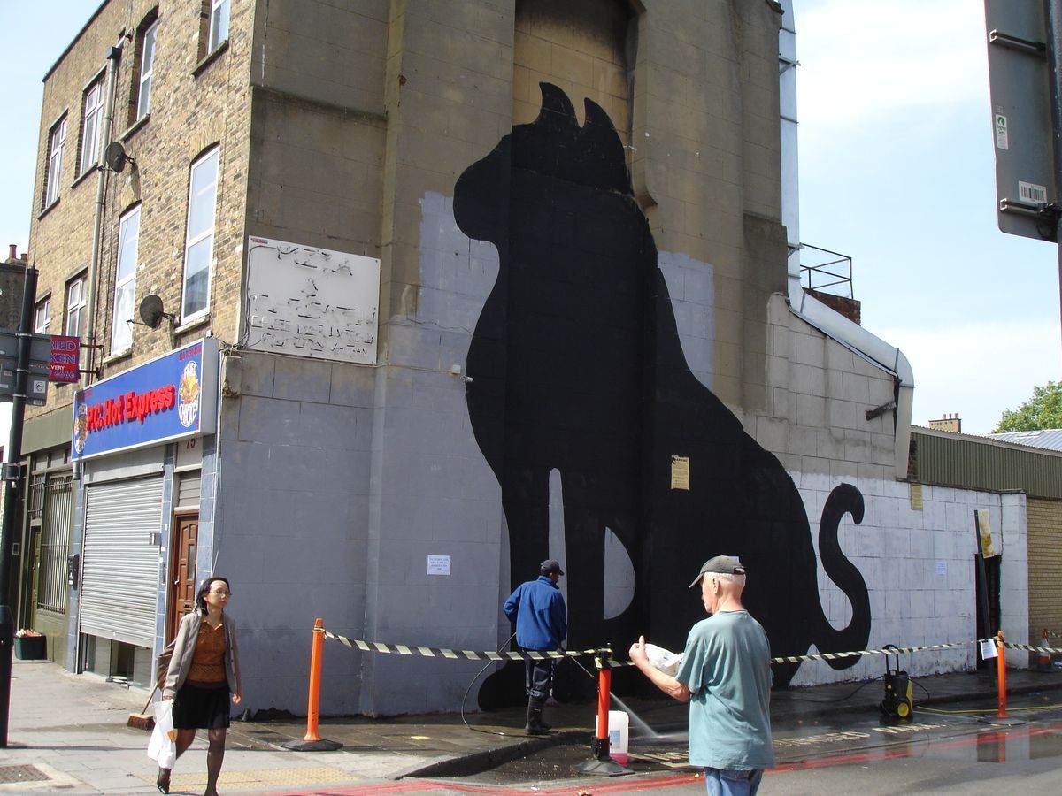 Giant black silhouette of a sitting cat painted across a building corner, workers pressure-washing the wall below, a pedestrian and shop signage in front.