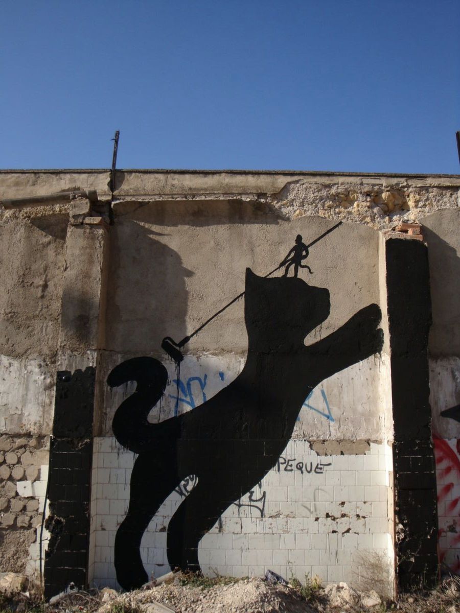 Black silhouette of a leaping cat with a tiny figure standing on its head holding a long paint roller pole, painted on a weathered concrete and tiled wall.