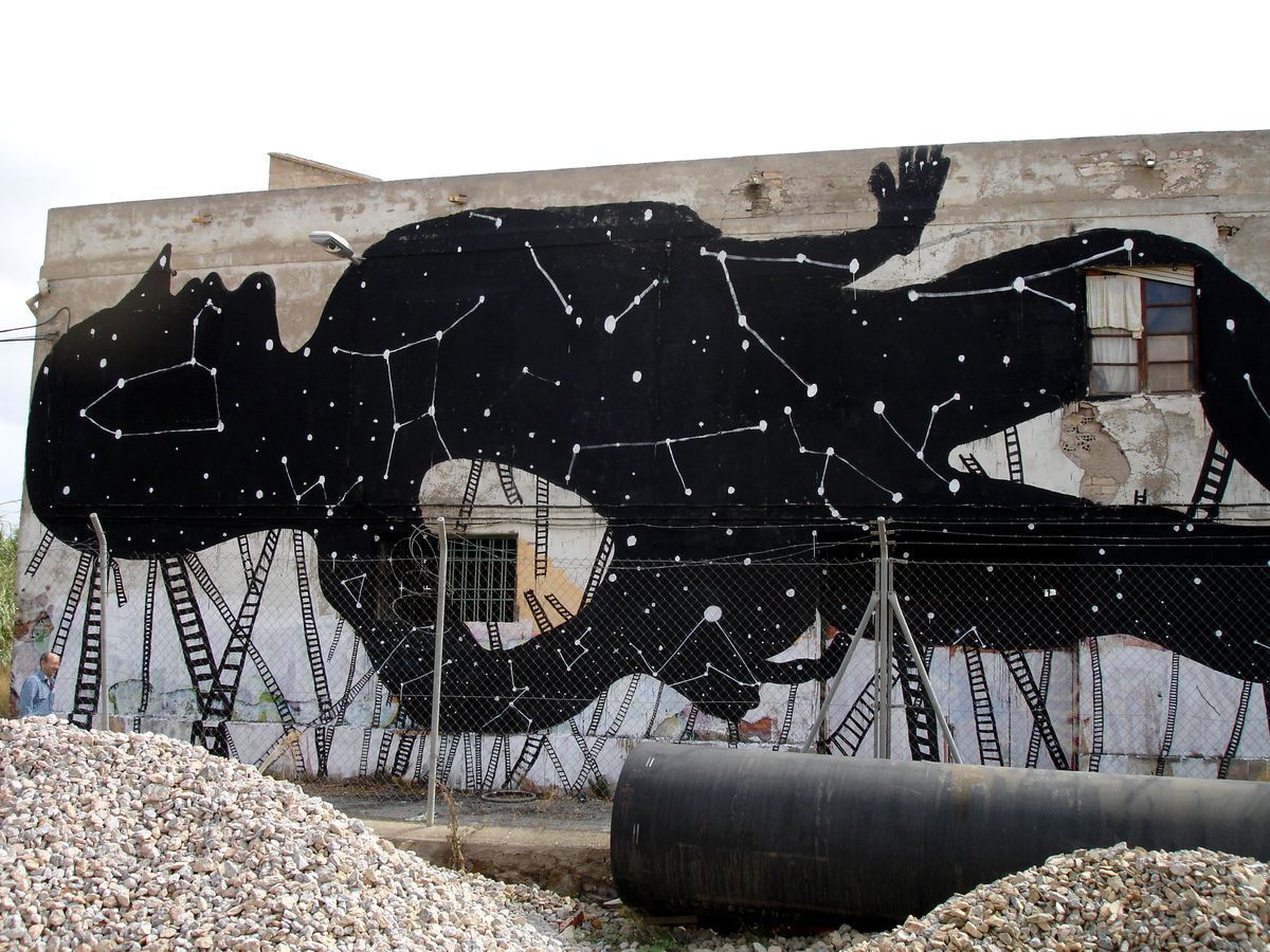 Wide view of a black silhouette animal covered in constellation patterns, a hand visible at the upper edge, ladders and a chain-link fence below, gravel piles in the foreground.