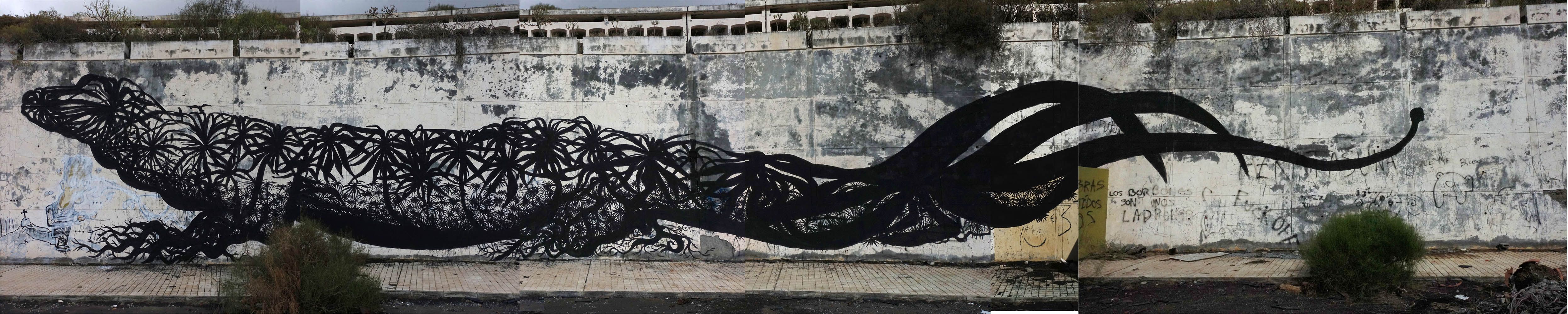 Extremely elongated black silhouette mural of a lizard-like creature stretched across a long panoramic wall, its body densely textured with an intricate pattern of radiating leaf and flower shapes, painted along a weathered concrete embankment.