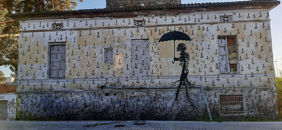 Wide view of a building facade entirely covered in a repeating small anchor pattern, with a black silhouette mural of a tree-like humanoid figure holding an open umbrella painted over the lower half.