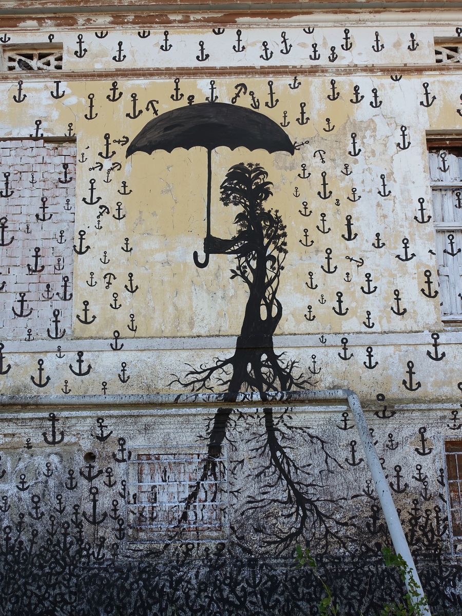 Black mural on a wall covered in a repeating small anchor pattern, depicting a tree-like humanoid figure with a leafy crown holding a large open umbrella, its roots reflected below a horizontal drainpipe.