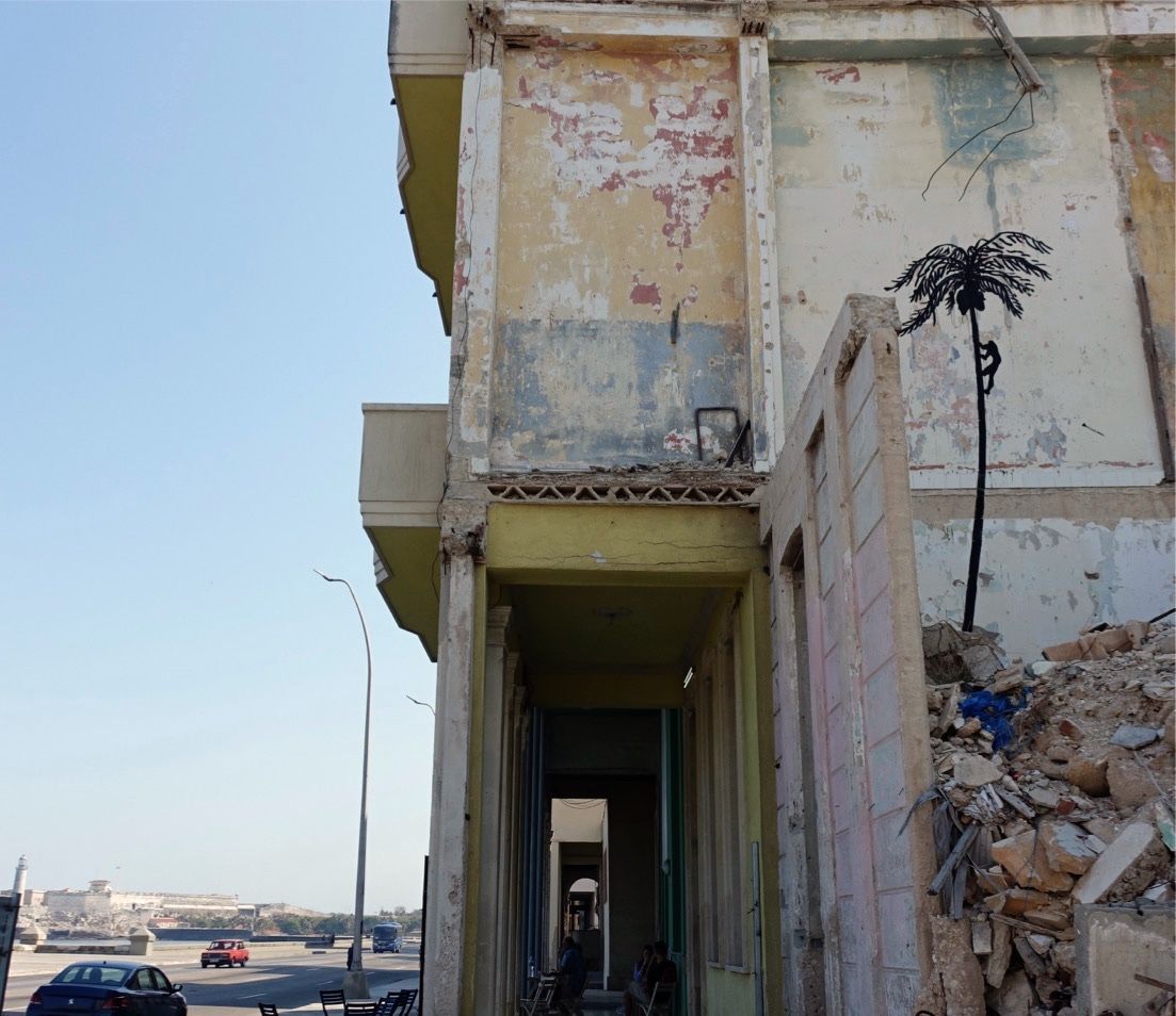 Small black silhouette mural of a palm tree with a person climbing it, painted on the weathered upper wall of a building beside a rubble pile, with a coastal road and fortress visible in the distance.