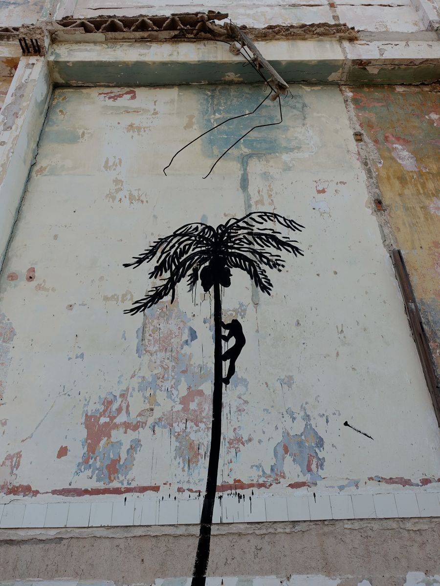 Close-up view of a black silhouette mural of a palm tree with a person climbing its trunk, painted on a heavily weathered, multicolored peeling wall.