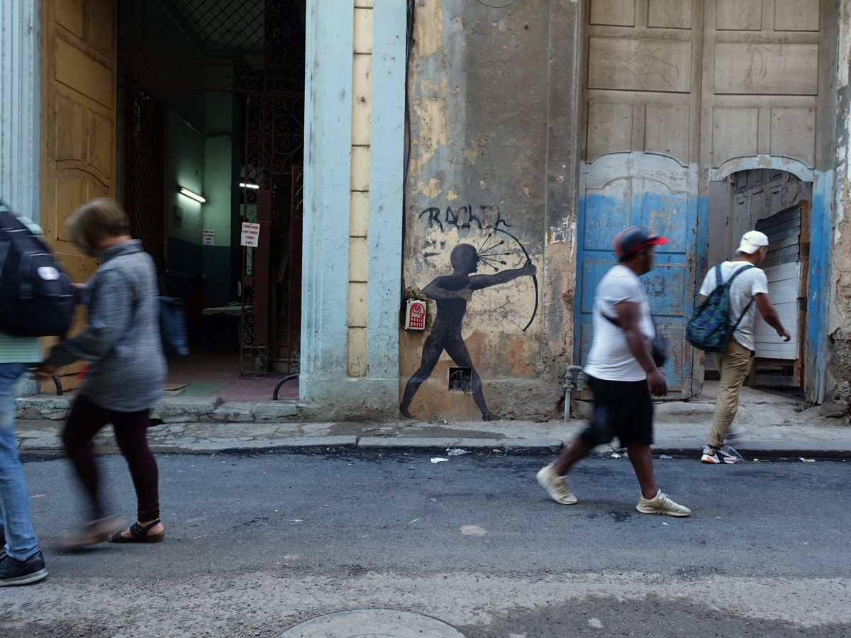 Black silhouette mural of a boy drawing a bow and arrow, painted on a weathered building facade above a doorway, with pedestrians walking past on the street below.