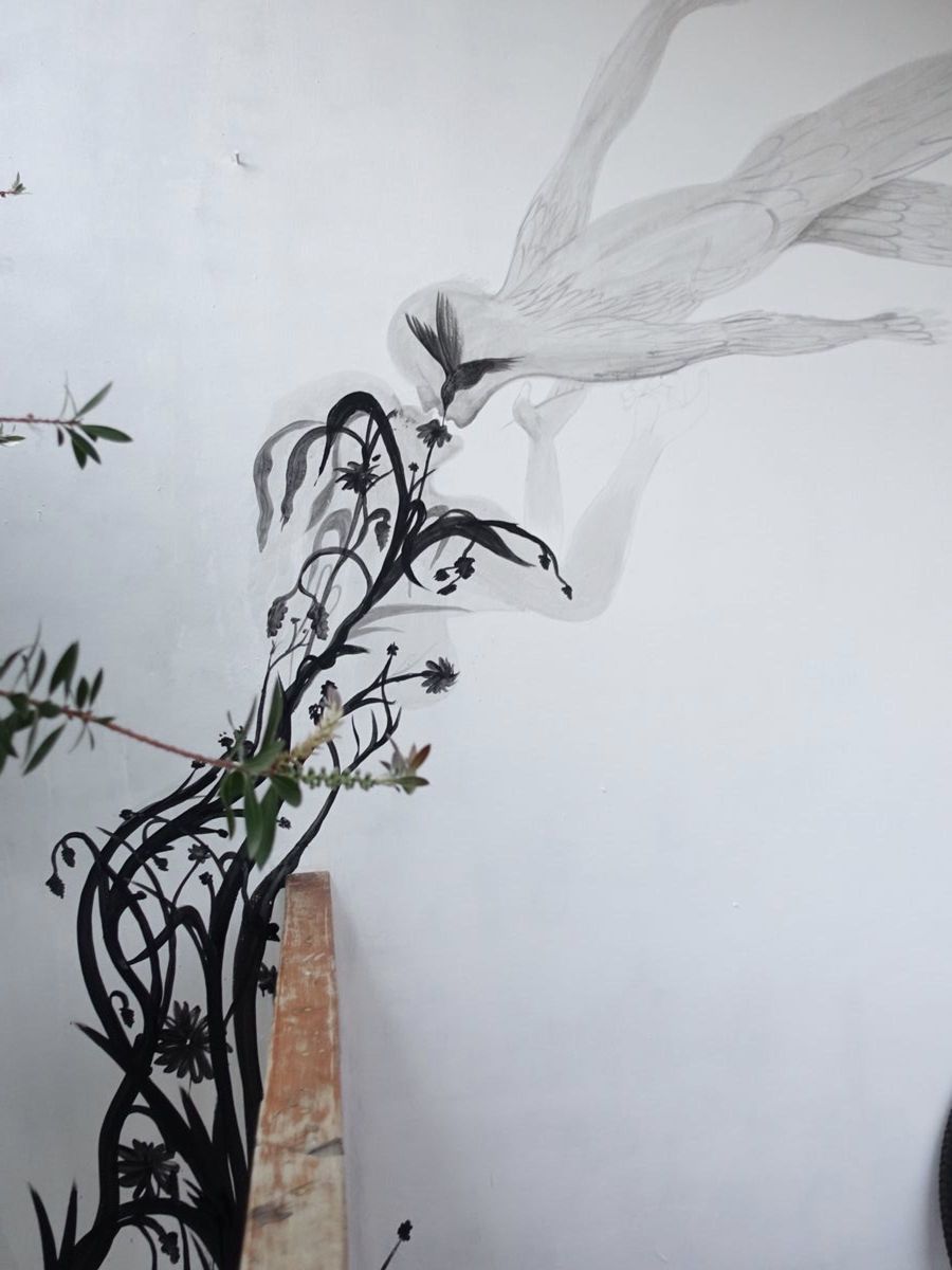 Close-up detail of a pale grey mural showing a bird's head reaching toward a black-painted flowering branch, next to a real potted plant placed against the wall.
