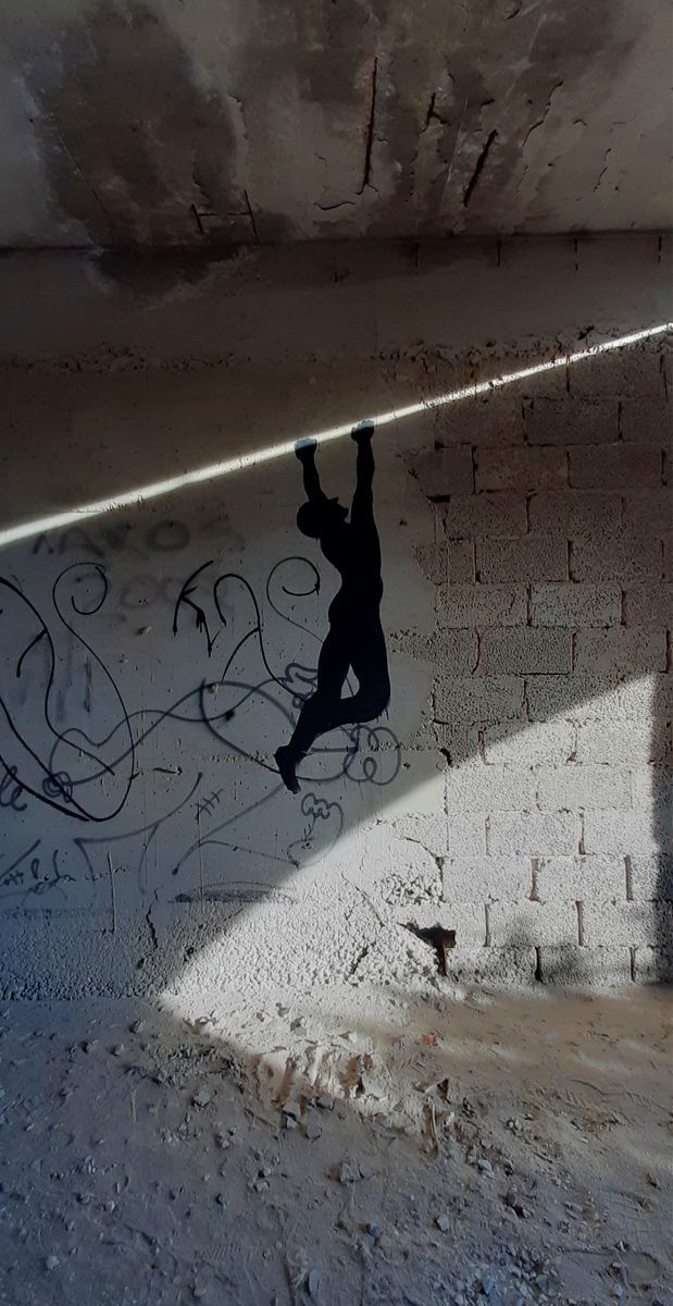 Black silhouette mural of a figure hanging by its hands from an overhead beam, legs bent, painted on a concrete wall covered in scribbled graffiti, lit by a diagonal beam of sunlight.