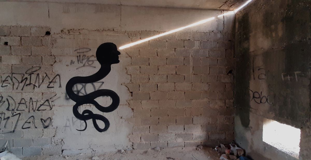 Black silhouette mural of a human head atop a long looping snake-like body, painted on a cinder block wall next to a shaft of light entering through a gap in the ceiling.