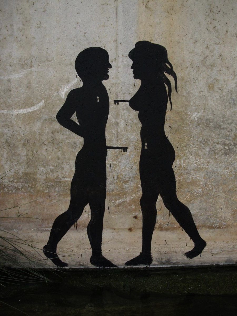 Black silhouette mural of a nude man and woman facing each other, each with a keyhole on their torso and holding a key extended toward the other's chest, painted on a weathered concrete wall.