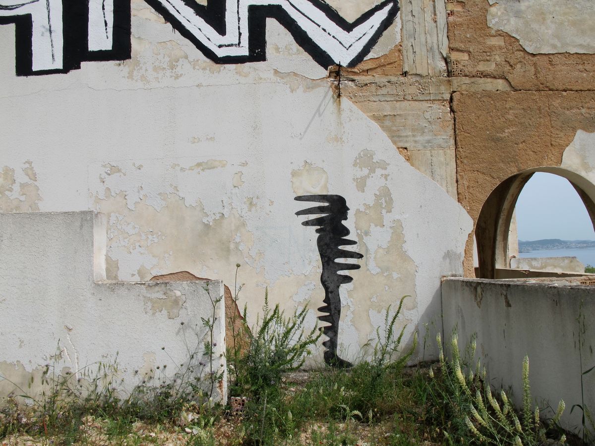 Small black silhouette mural of a wavy, zigzagging human figure painted on a weathered stone wall near a stone archway overlooking the sea, beneath a black-and-white graffiti tag.