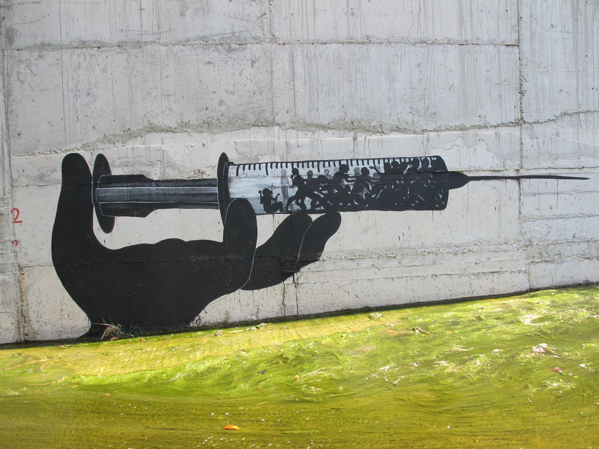 Black silhouette mural of a hand holding a large syringe, with a crowd of running figures depicted inside the syringe barrel, painted on a concrete wall above a green algae-covered canal.