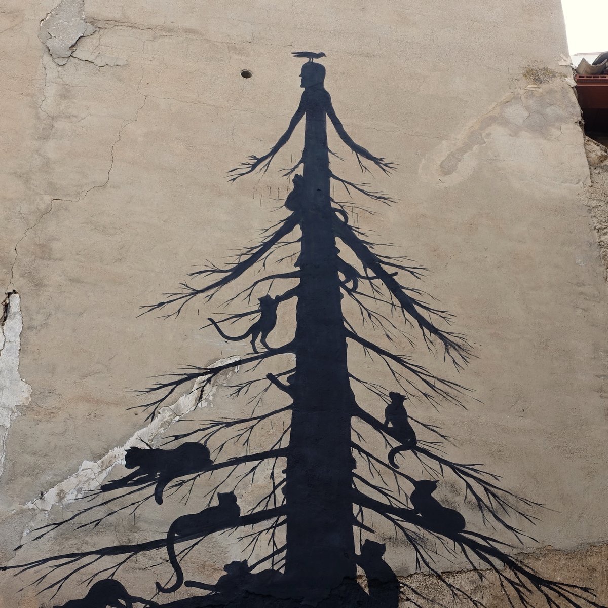 Black silhouette mural of a tall bare tree topped with a human head and outstretched branch-arms, a crow perched on its head, with several cats climbing along the lower branches, painted on a weathered plaster wall.