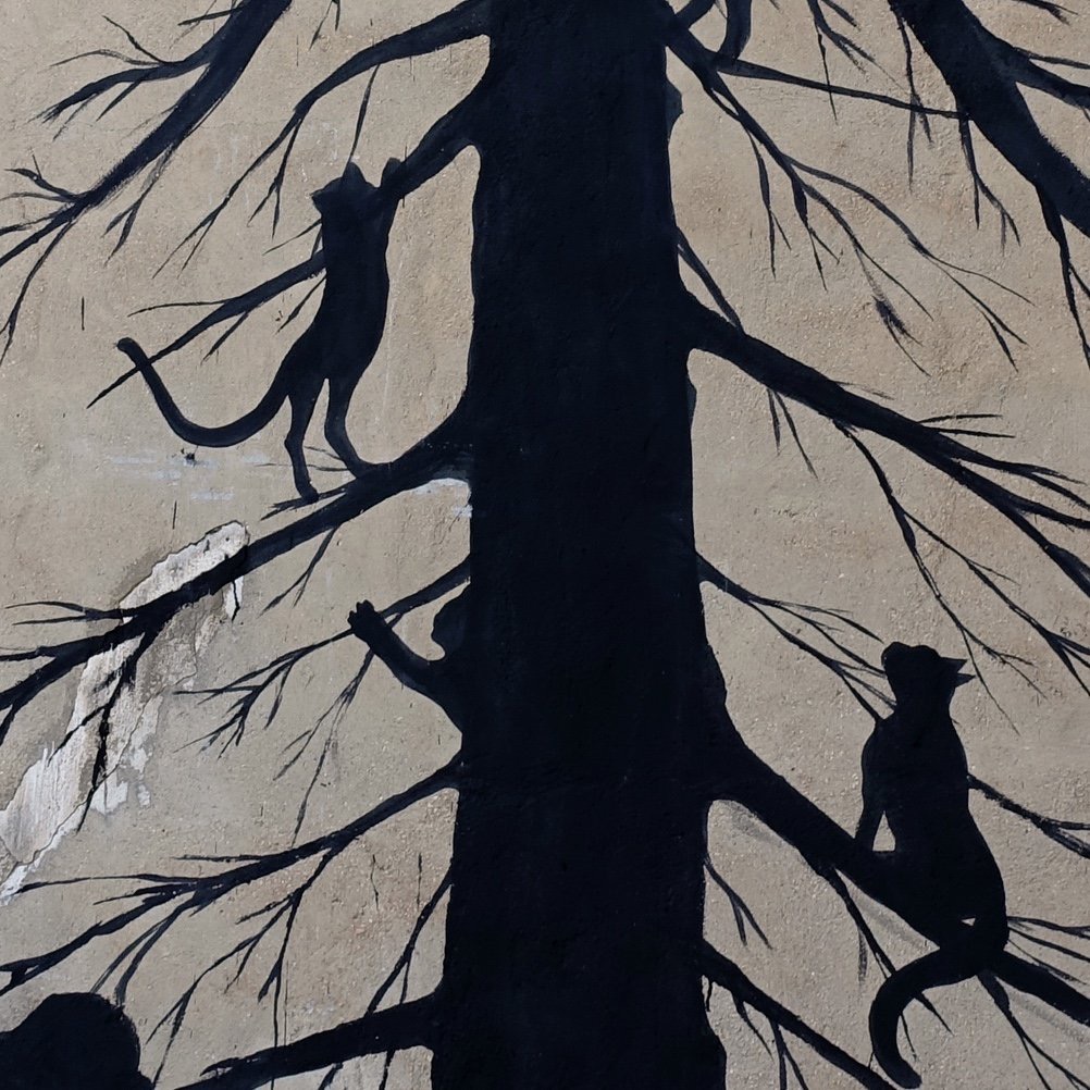 Close-up detail of a black silhouette mural showing bare tree branches with two cats climbing among them, against a rough concrete wall.