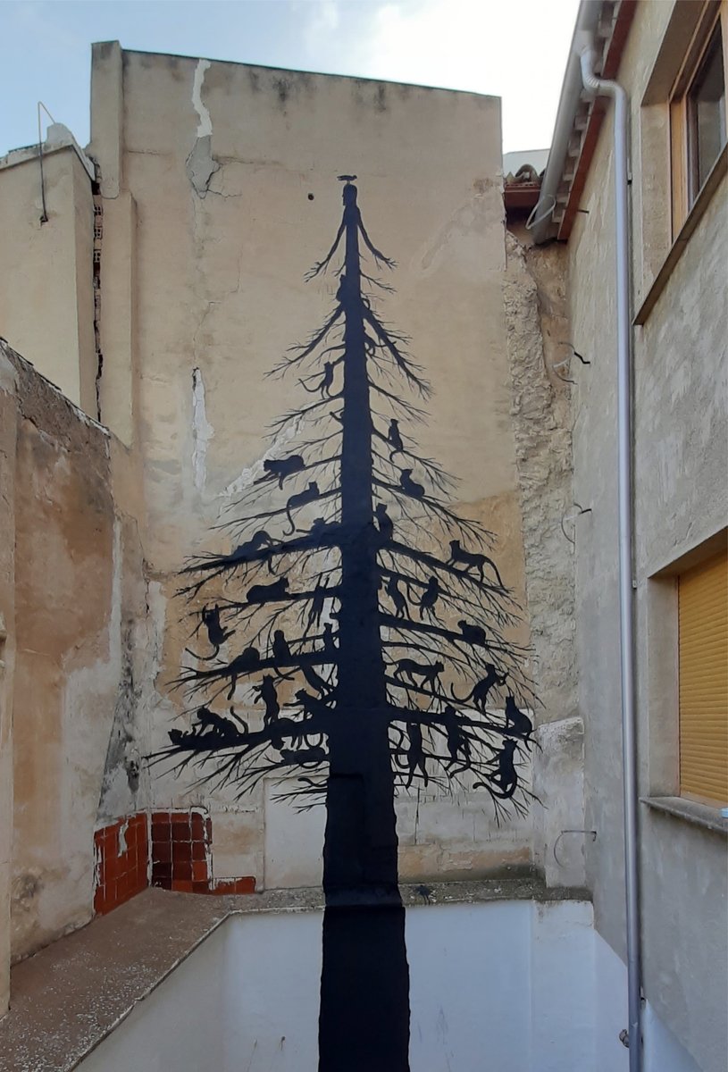 Full wide view of a tall black silhouette tree mural covering a building wall between two structures, densely populated with numerous climbing cats along its branches, a bird at the very top.