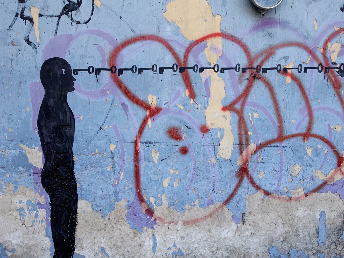 Black silhouette mural of a standing human figure with a keyhole cut into its head, facing a horizontal row of small keys, against a weathered blue wall covered in red and purple spray-paint circles.