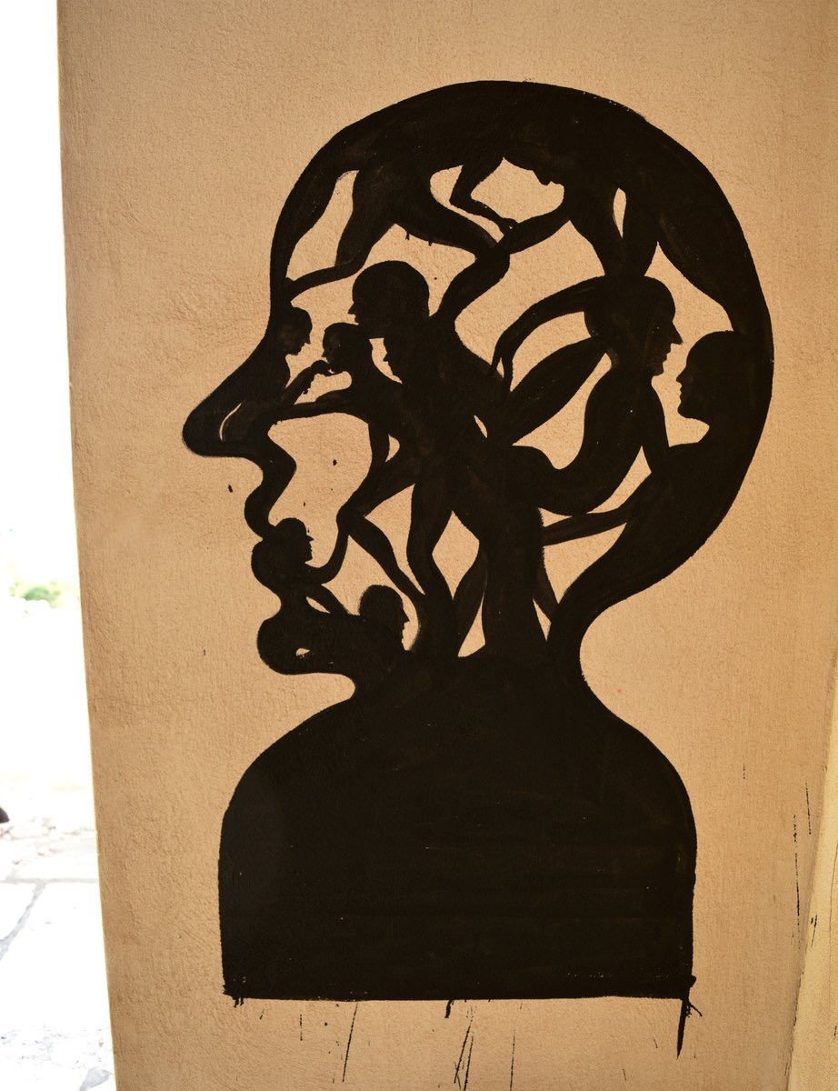 Black mural silhouette of a human head in profile on a beige wall, its interior filled with intertwined nude human figures and branch-like forms resembling a tree.