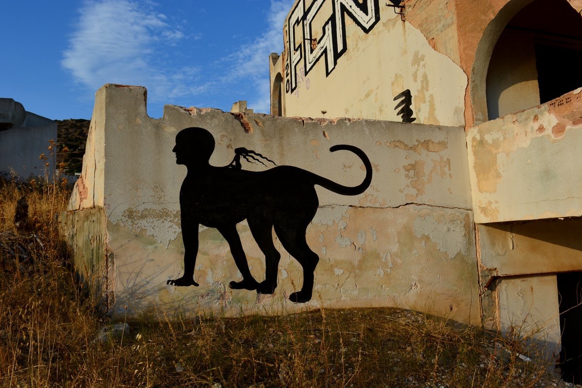 Black silhouette street art mural of a sphinx, a human head on a four-legged animal body with a long curved tail, painted on the weathered plaster wall of an abandoned building.