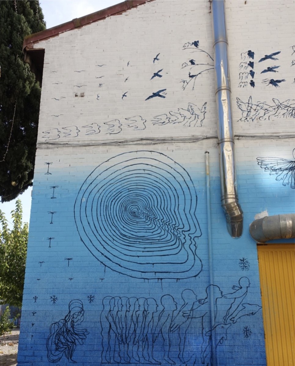 Large-scale wall painting split into a white upper section with fine-line birds in flight and a blue lower section, centered on a spiral tree-ring pattern that resolves into a human face in profile, with a row of walking silhouetted figures along the bottom edge.