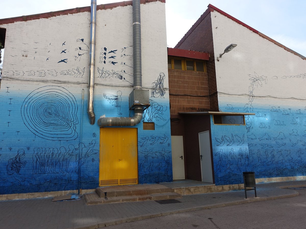 Wide view of a two-story building facade covered in a large mural: white sky section with birds and vines above, blue lower section with a tree-ring face motif and a line of walking figures, a yellow metal door built into the composition.