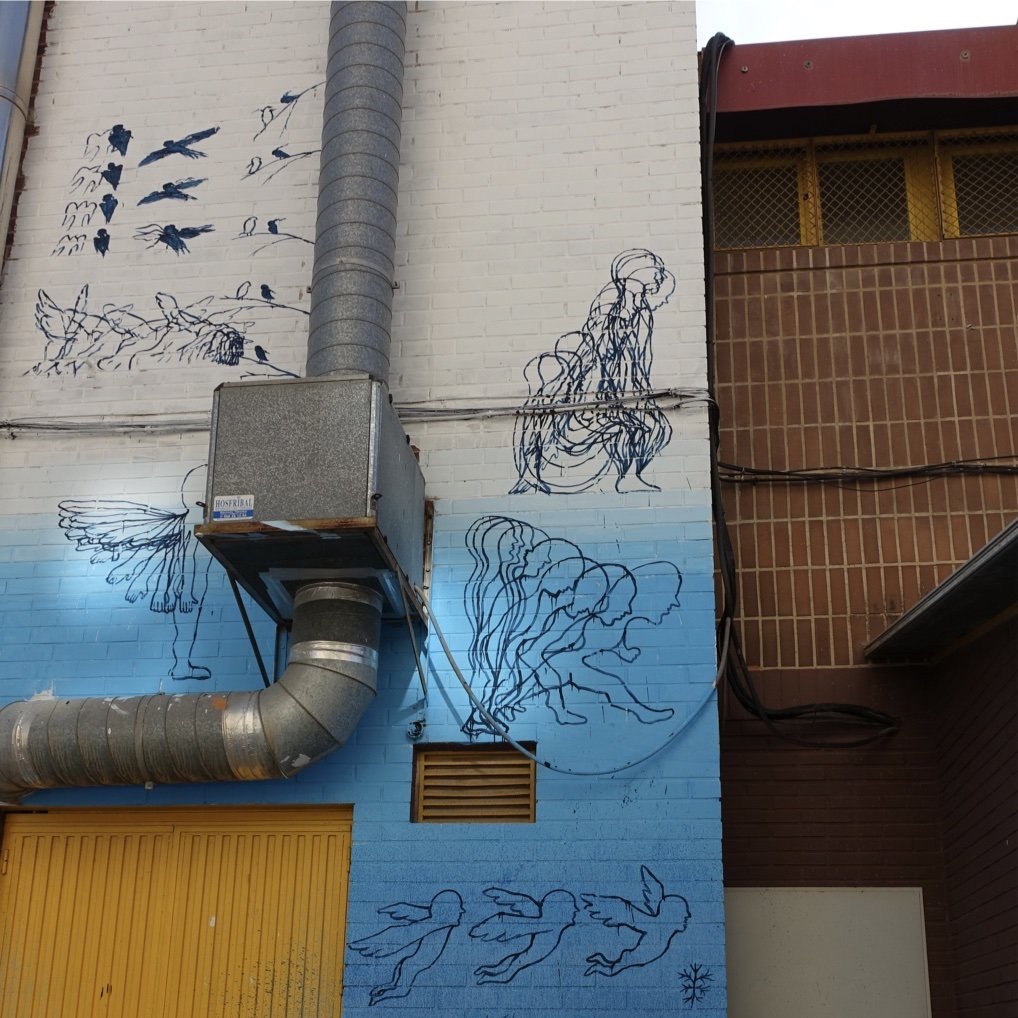 Detail of a black-and-white mural showing flying birds, a seated figure drawn in fine outline, a chain of walking silhouettes, and small outlined angels near a yellow door and exposed ventilation pipes.