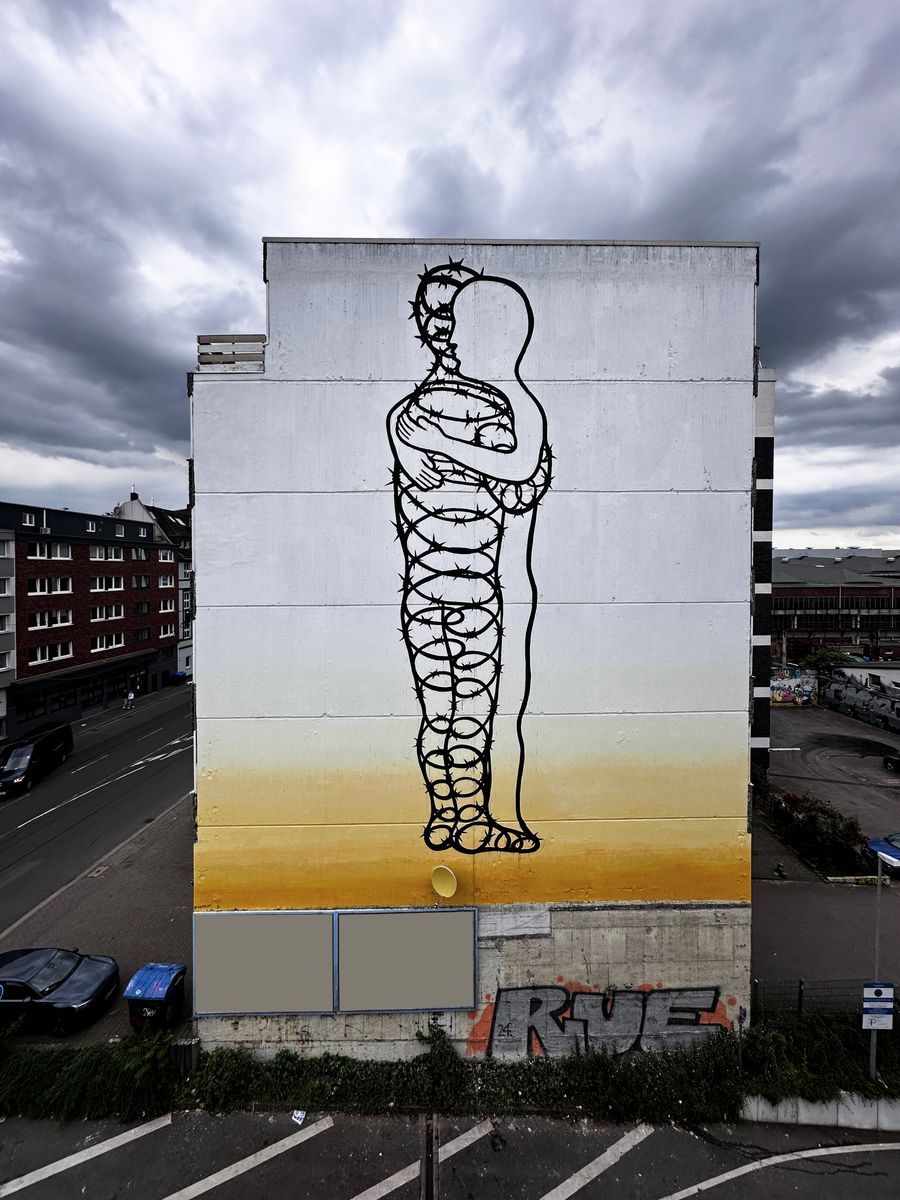 Tall building facade mural of a black outline figure wrapped head to foot in coiled barbed wire, arms crossed over the chest, painted on a white wall fading to yellow at the base, graffiti tag below.