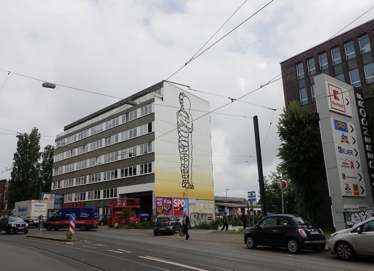 Wide street view of the same barbed-wire figure mural on a building corner beside a Kaufland shopping center sign, tram wires crossing overhead and cars parked below.