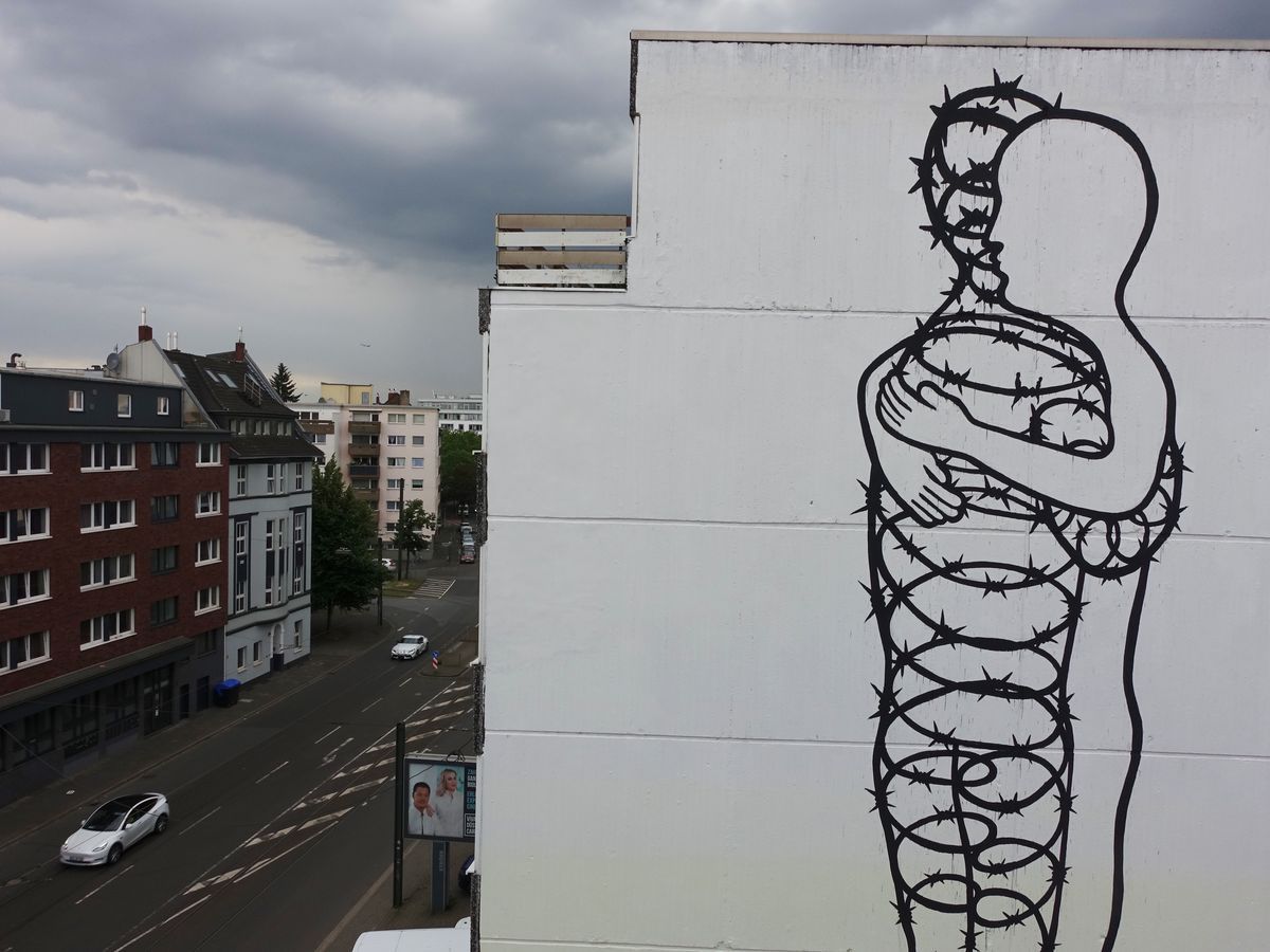 Aerial view looking down at the barbed-wire figure mural on the building's white facade, city street and rooftops visible below.