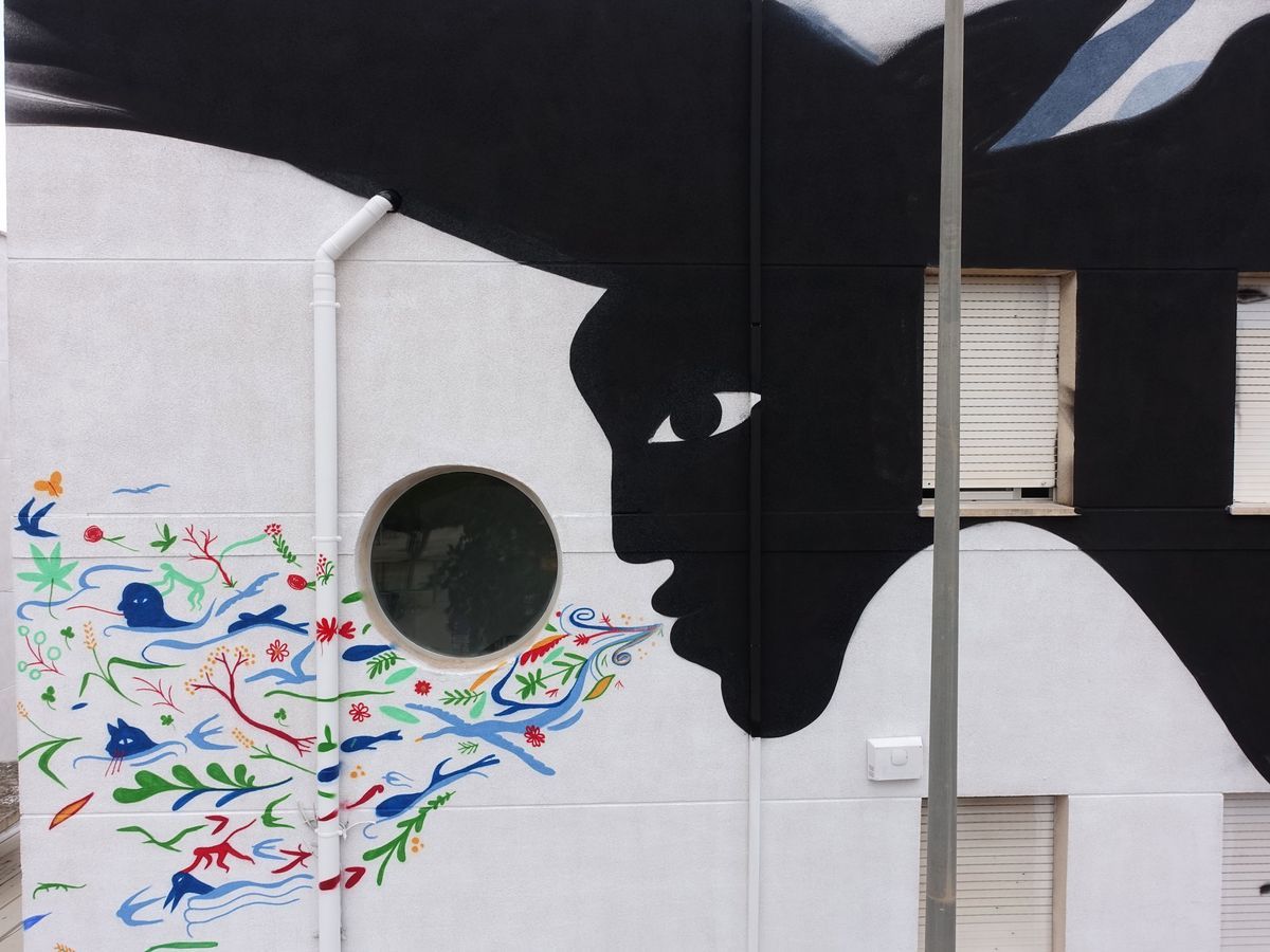 Mural of a black silhouetted profile face pressed close to a round window, a burst of colorful painted flora and fauna (birds, fish, flowers) flowing from its mouth across the white wall.