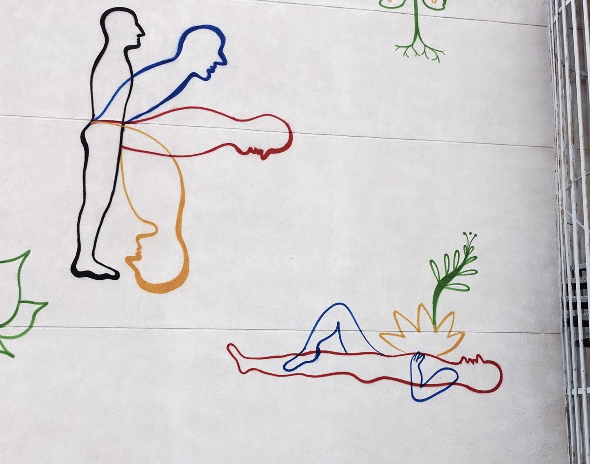 Line-art mural of overlapping colored outline figures in black, blue, red, and orange, one standing figure and one reclining figure, with painted leaves and a flower sprouting nearby.
