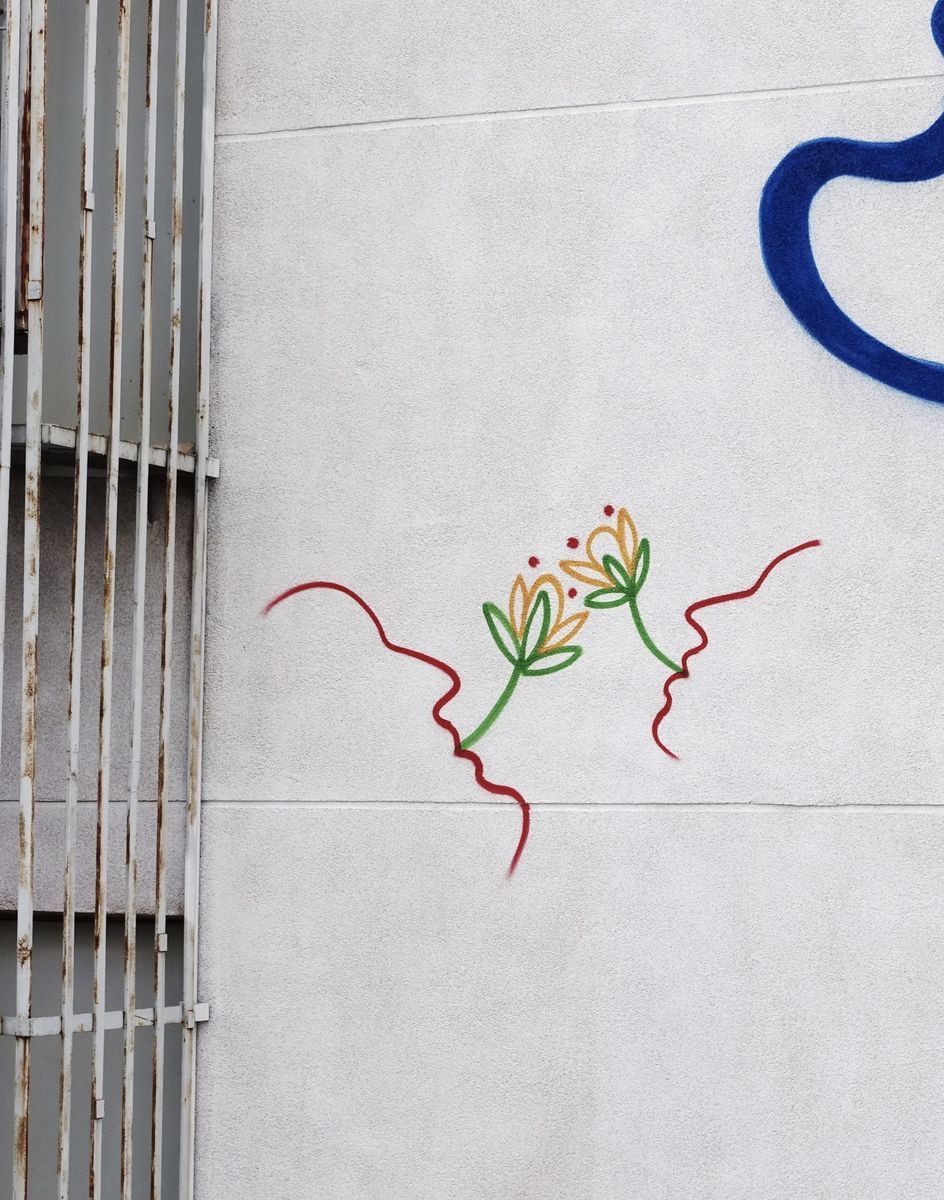 Small painted detail of two red wavy line profiles facing each other with a green and orange flower blooming between them, on a plain wall beside a rusted metal grille.
