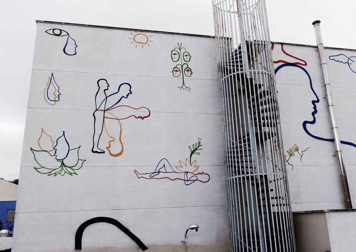 Wide view of a large multi-figure mural on a building corner, showing outline figures in blue, black, red, and orange intertwined with plants, faces, teardrops, a sun, and a spiral staircase running up the wall.