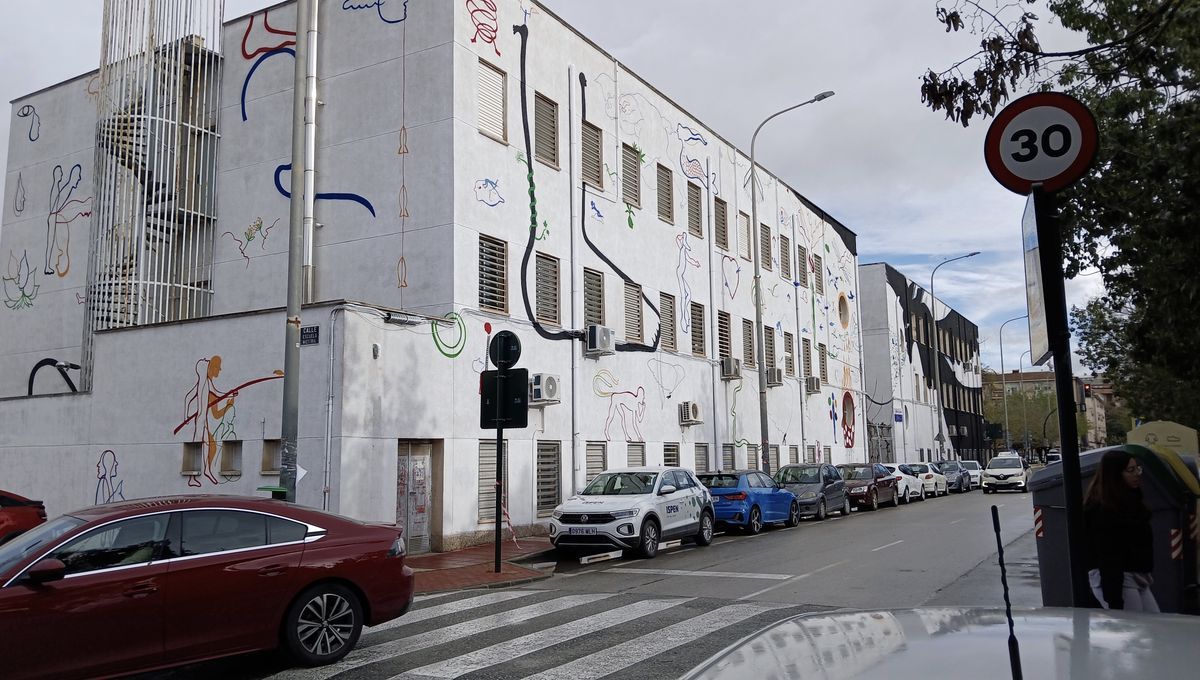 Wide street corner view of a multi-story building entirely covered in colorful outline mural figures and shapes, a spiral staircase and speed limit sign visible, cars parked along the street.