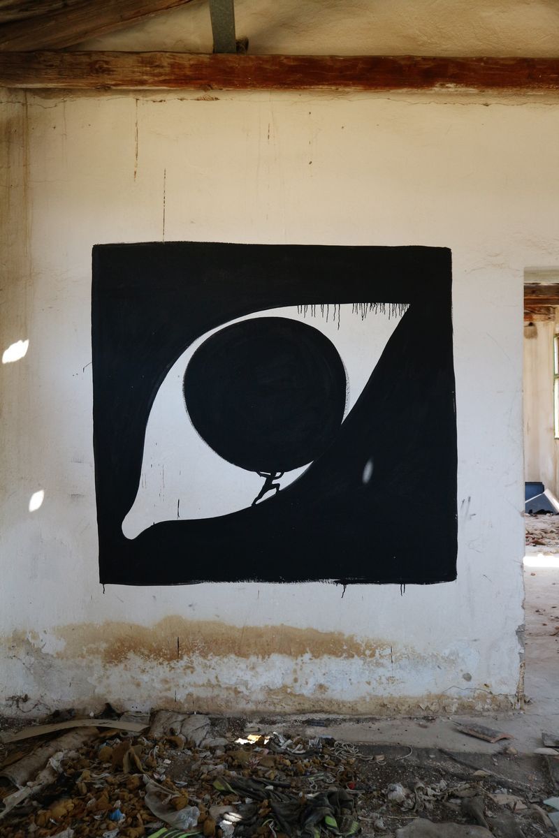 Black square mural of a large eye shape containing a round black boulder pushed by a tiny silhouetted figure at its base, painted inside a ruined stone building, debris scattered on the floor.