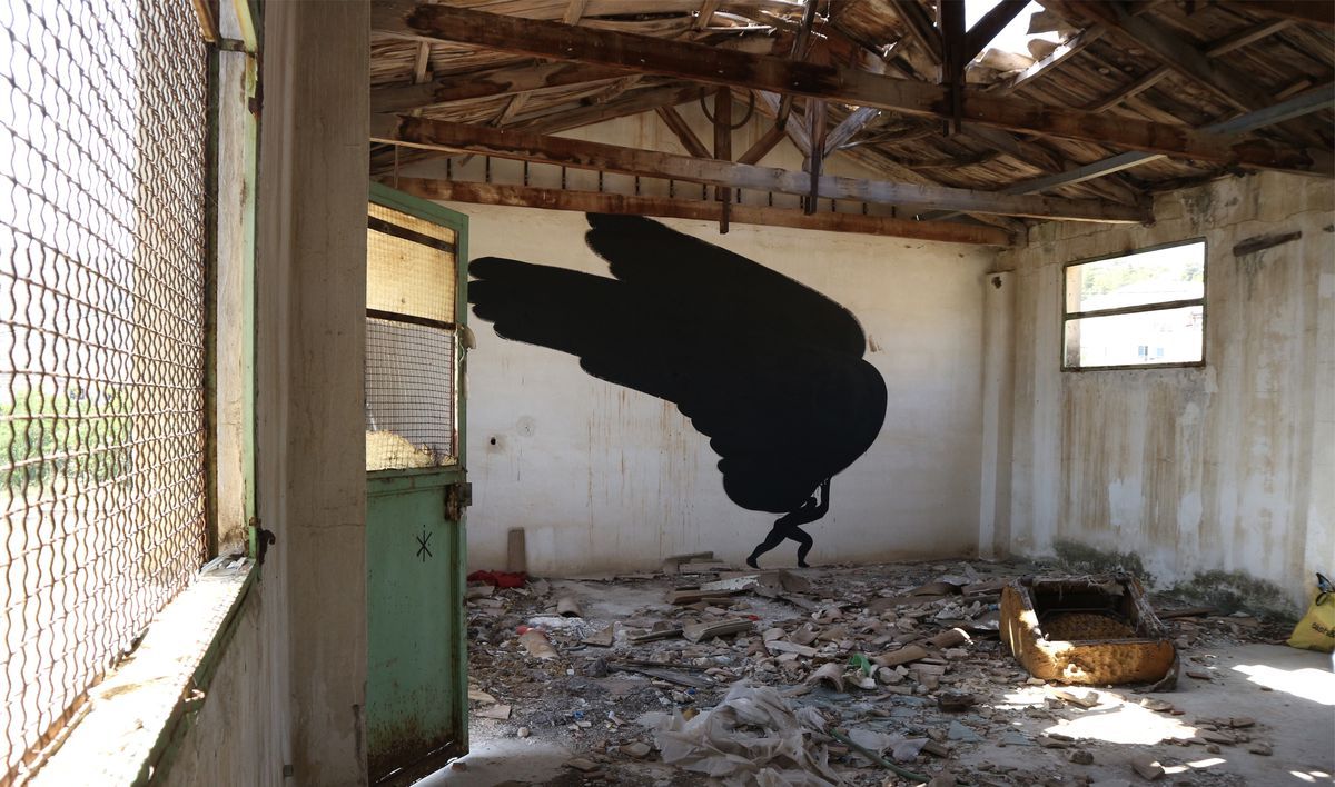 Interior of a roofless ruined building, a huge black wing-shaped mural on the back wall with a small silhouetted figure appearing to carry it, rubble and a green door frame in the room.