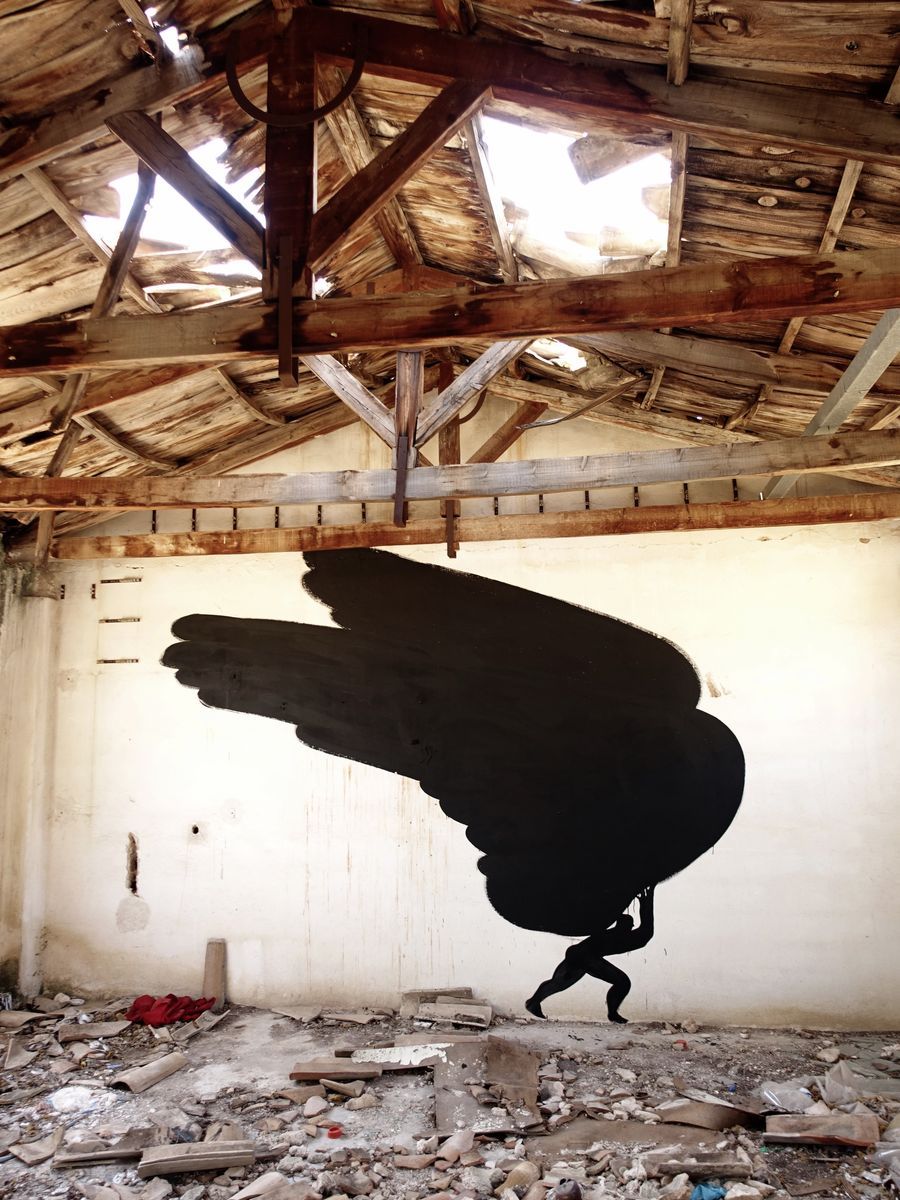 Closer view of the same black wing mural under exposed wooden roof beams with skylight gaps, a tiny figure carrying the wing at its base, debris on the floor.