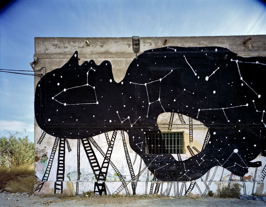 Black silhouette of an elongated animal figure covered in constellation patterns of stars and connecting lines, ladders leaning against the wall below it, a barred window set into the silhouette.