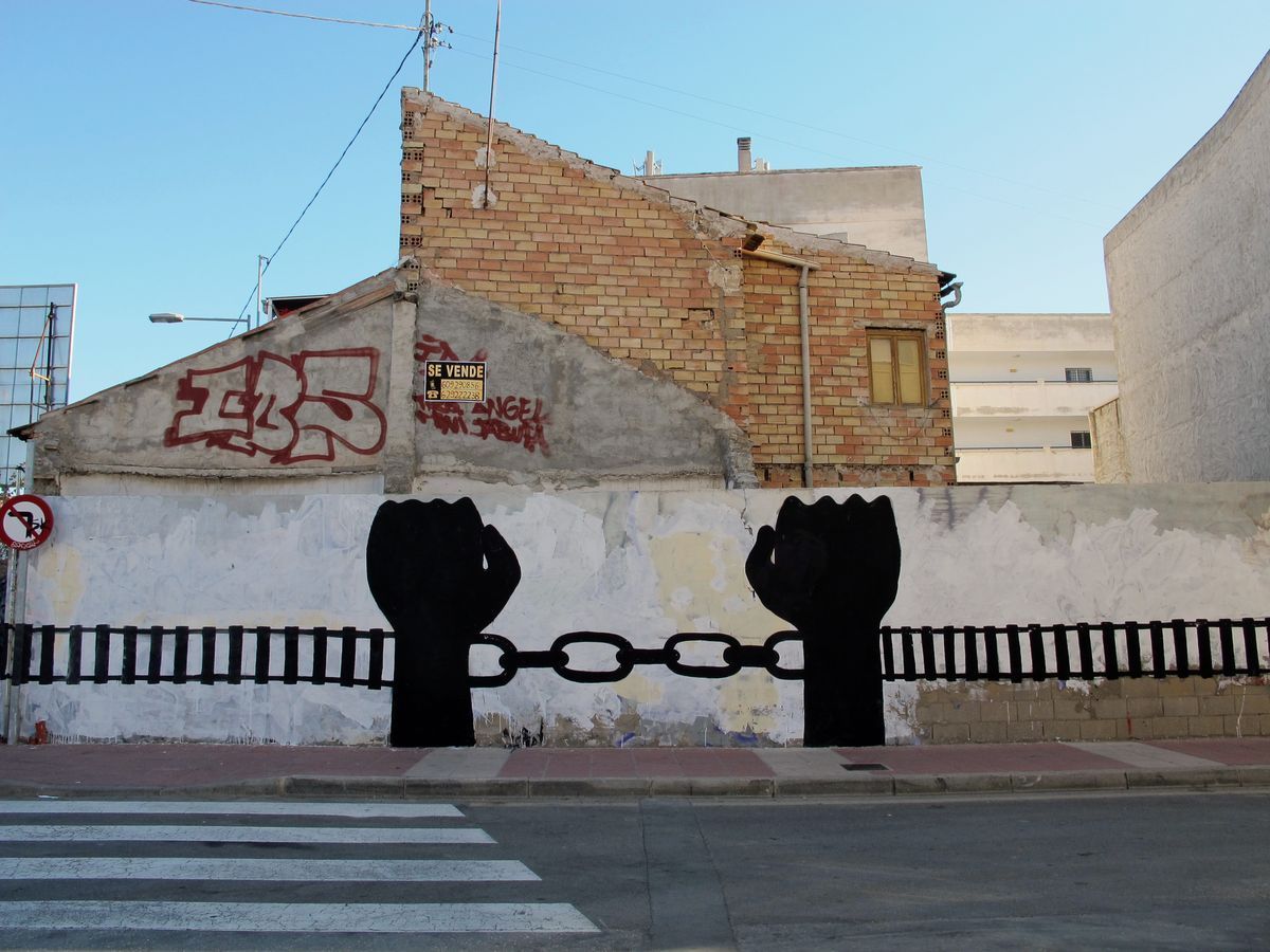 Two black silhouette raised fists connected by a chain, painted above a low wall topped with a rail-like fence, a crumbling brick gable and a no-parking sign visible behind, a crosswalk in front.