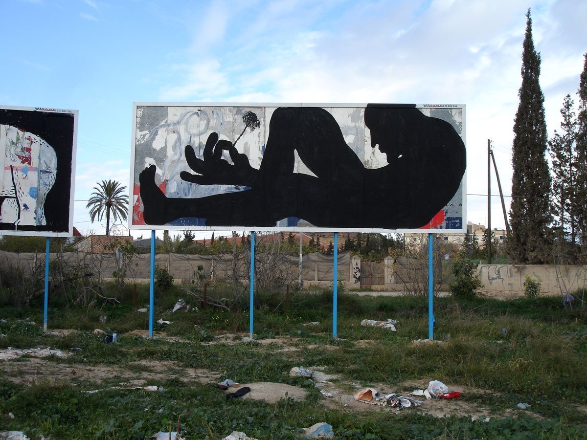 Giant reclining black silhouette figure smoking a cigarette, painted across a roadside billboard, palm trees and another billboard visible in the background.