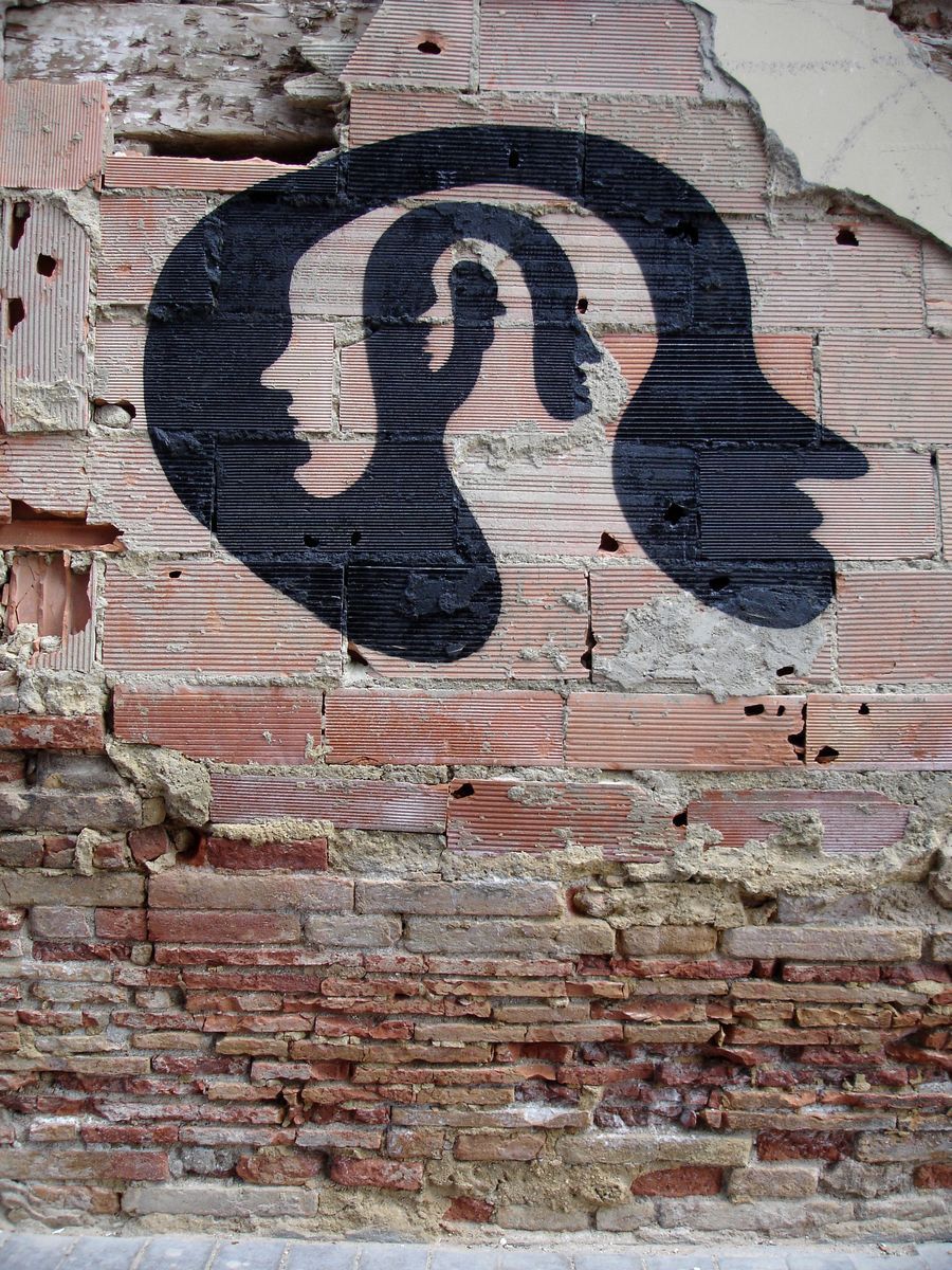 Black silhouette spiral shape formed by two mirrored human profile heads facing each other, painted on a weathered exposed brick wall.