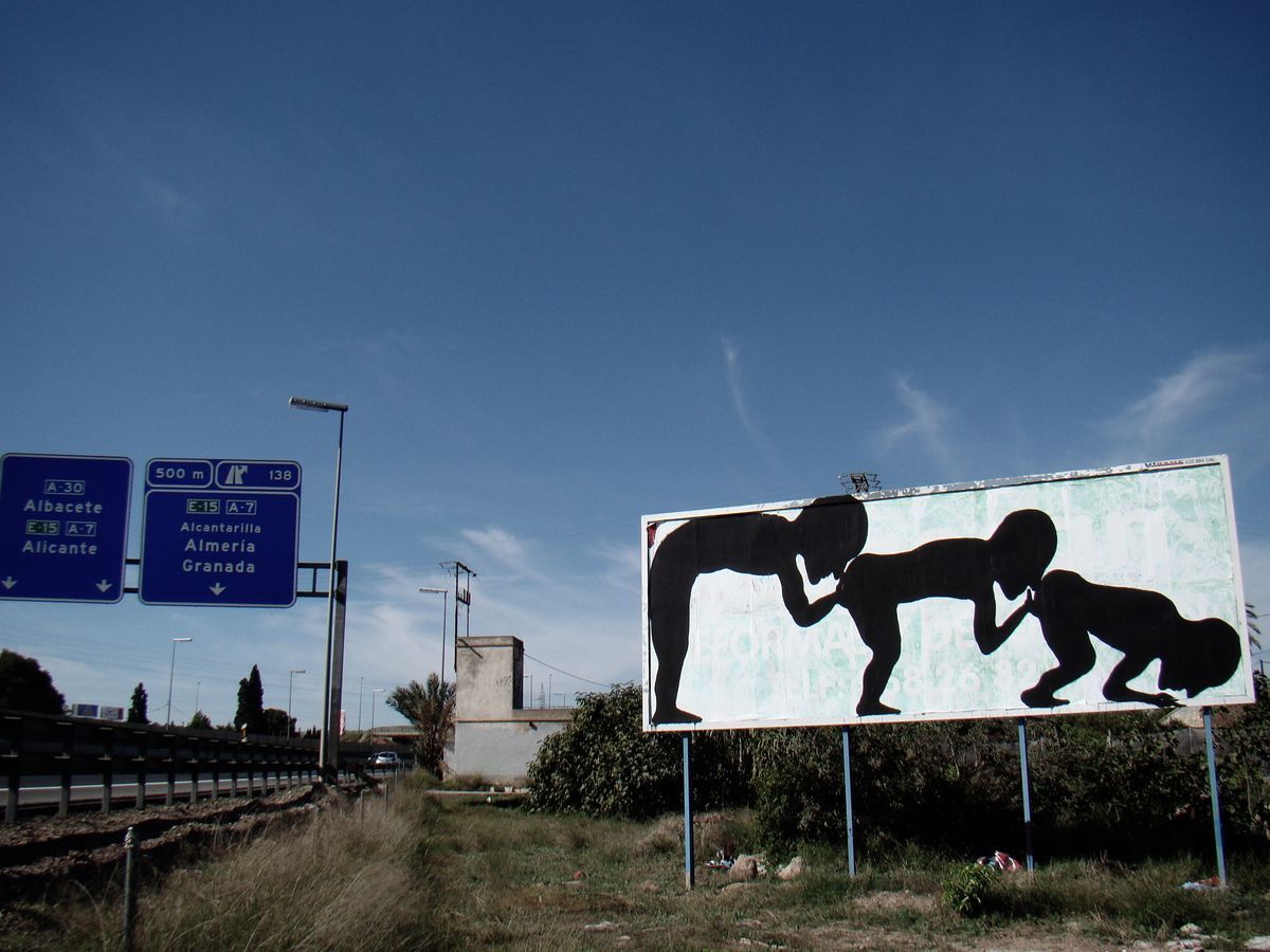Roadside billboard painted with three black silhouette figures in a row bent forward kissing the buttocks of the one in front, climbing over a partly visible advertisement, highway direction signs beside it.