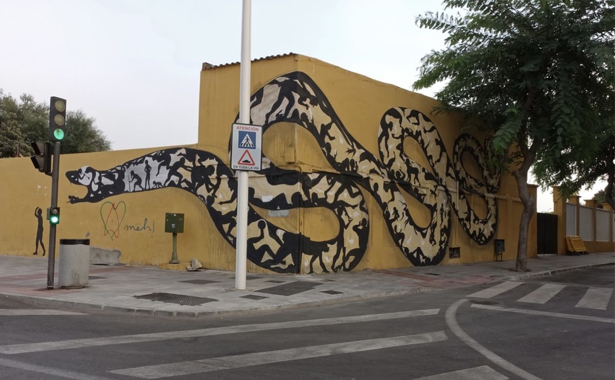Large black and beige mural of a snake winding across a building's corner and side walls, its body filled with silhouetted human and animal figures, painted at a street intersection with a traffic light and pedestrian crossing sign.