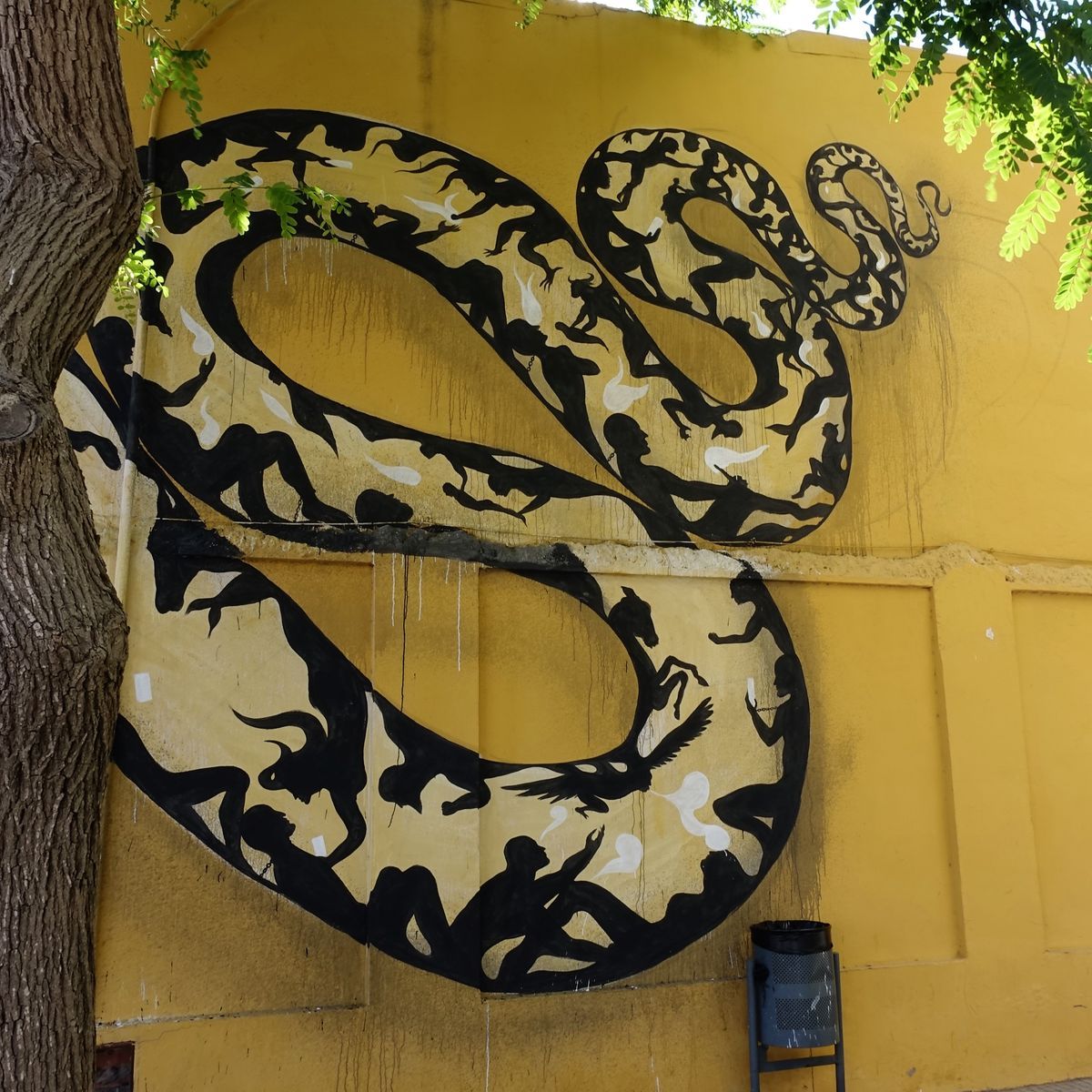 Closer view of the coiled snake mural on a yellow wall, its looping body packed with black silhouettes of people and animals, a tree and litter bin in front.