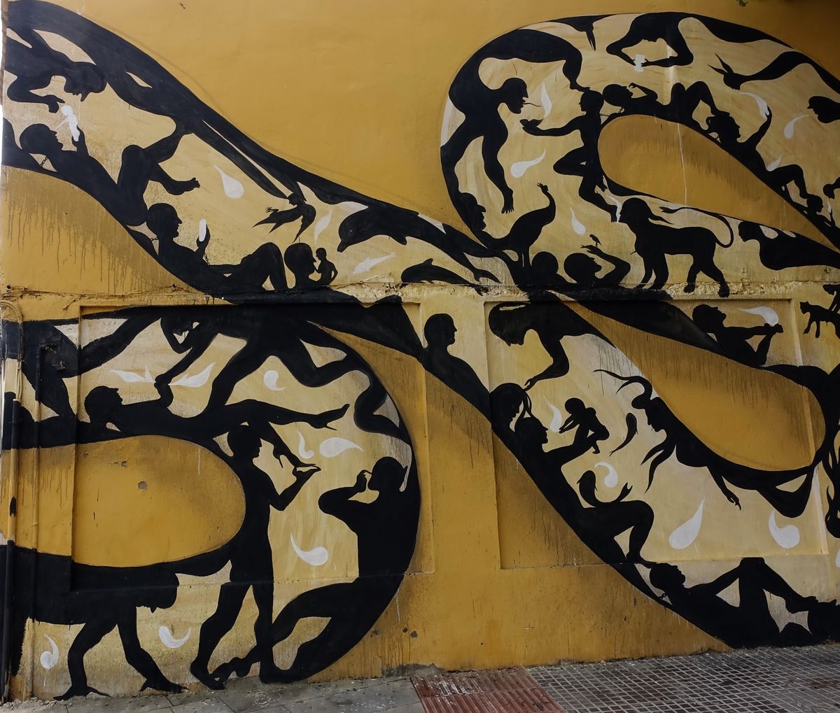 Close view of two coiled loops of the snake mural, densely filled with black silhouettes of people, a dolphin, a bird, and a monkey against the yellow wall.
