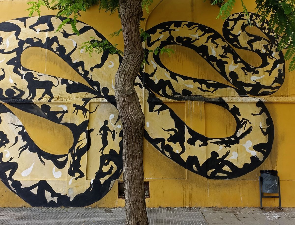 Wide view of two large coiled loops of the snake mural wrapping around a tree trunk, filled with black silhouettes of people and animals on the yellow building wall, a litter bin beside it.