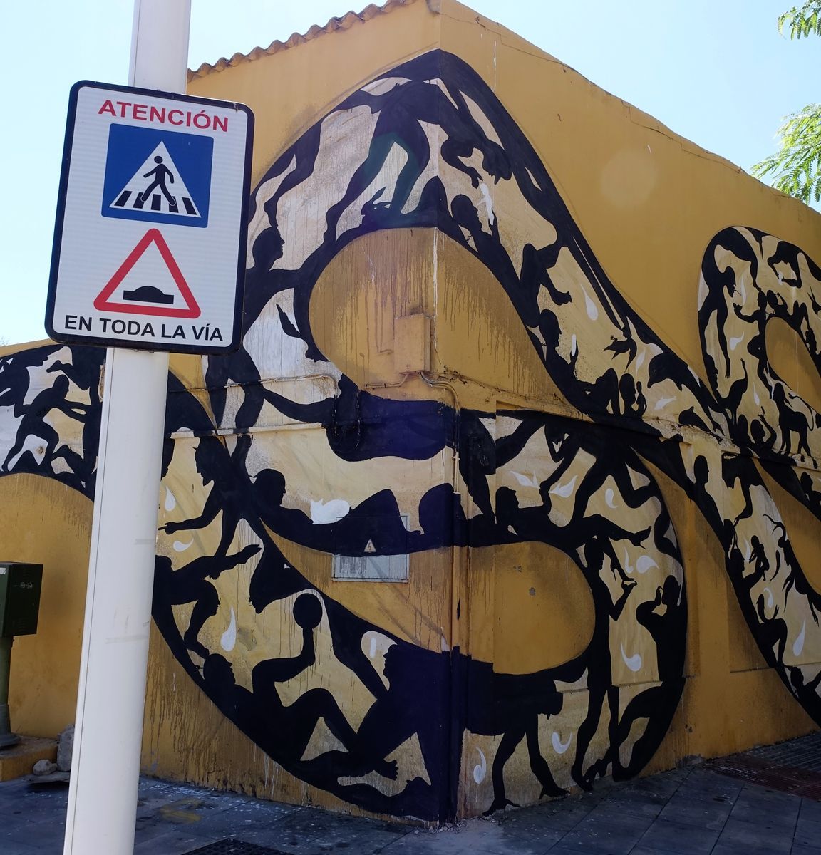 Corner view of the snake mural's coiled body filled with silhouetted figures climbing and playing, a pedestrian crossing and speed bump warning sign on a pole in front.
