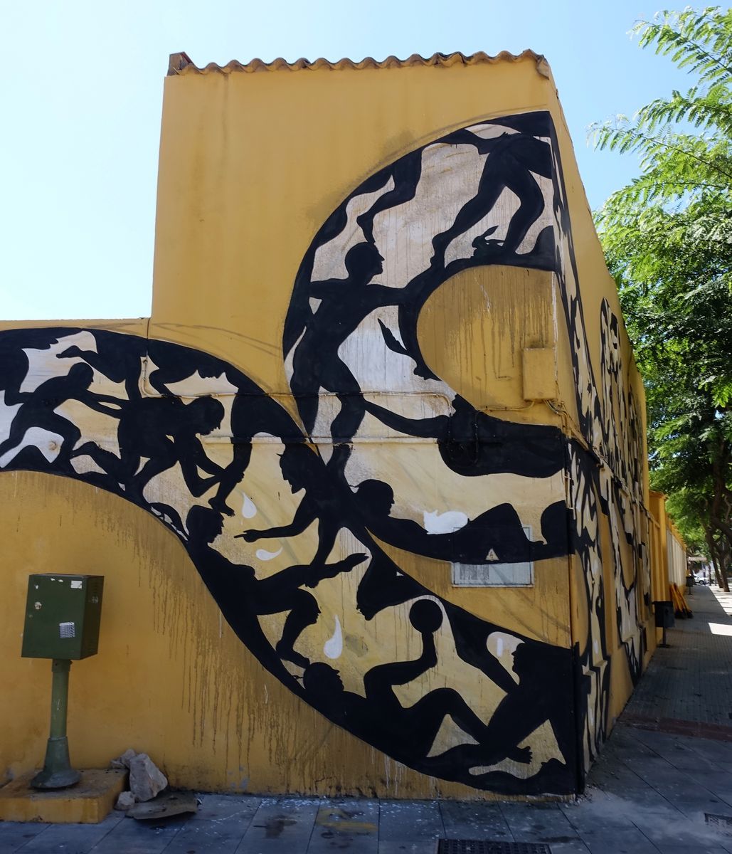 Side view of a looping section of the snake mural on a yellow wall, silhouettes throwing and holding a ball among the coils, a green utility box below.