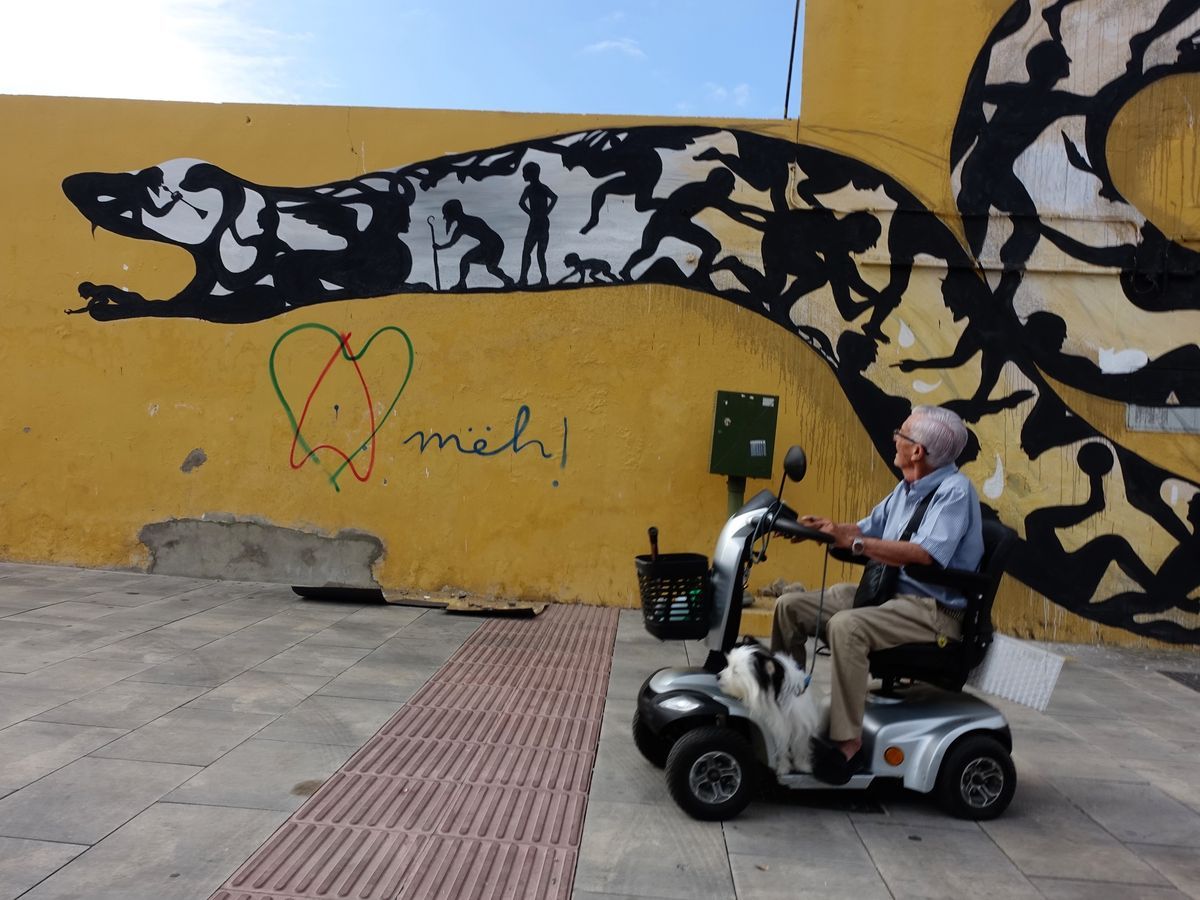 Wide view of the snake's open-mouthed head, its body filled with silhouettes including a child playing trumpet and an elderly figure with a cane, an elderly man on a mobility scooter with a dog passing in front, a green and red heart graffiti with "meh!" nearby.
