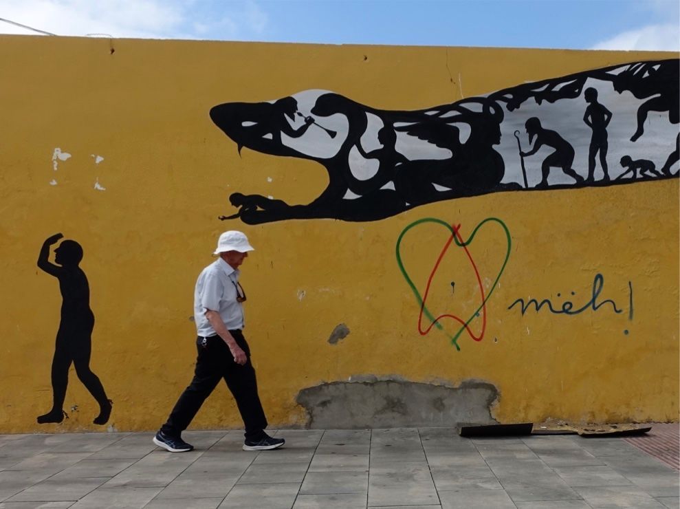 Wide view of the snake's head and neck on the yellow wall, an elderly man in a white hat walking past, a separate small walking silhouette figure to the left, red and green heart graffiti with "meh!" text.
