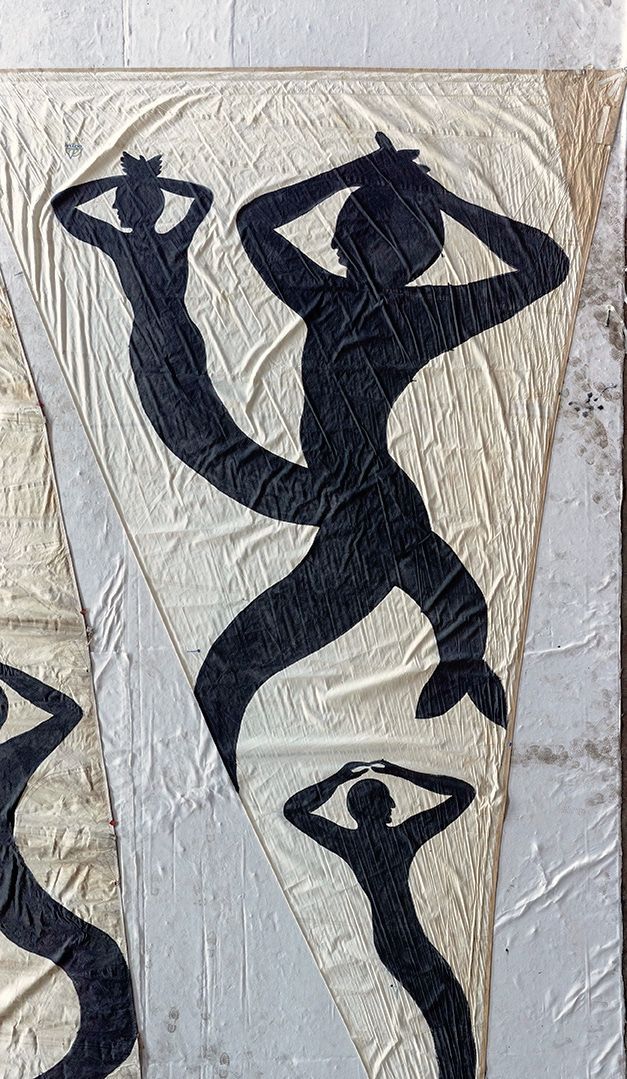 Close-up of a triangular sail with black silhouette figures of mermaids with raised arms and crowned heads painted on canvas, stains visible on the fabric.