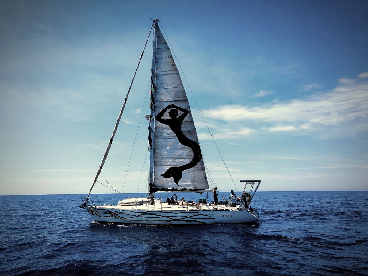 A sailboat at sea with its mainsail raised, displaying a black silhouette of a mermaid with raised arms painted on the white sail.